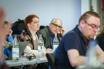 Eine männlich lesbare Person sitzt und schreibt, im Hintergrund weitere Personen, die zuhören