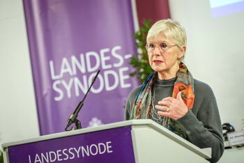 Eine weiblich lesbare Person mit Brille und blonden HAaren steht an einem Pult und spricht in eine Mikrofon.