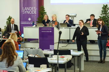 Eine Gruppe aus männlich und weiblich lesbaren Personen steht lachend um ein Pult vor einem langen Tisch, an dem weitere Personen sitzen und lächeln.