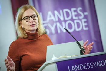 Eine weiblich lesbare Person mit Brille und einem Rollkragenpulliver steht an einem Pult und spricht.