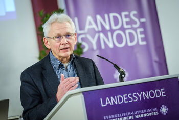 Eine männlich lesbare Person mit Brille und weissem Haar steht an einem Pult uns spricht.