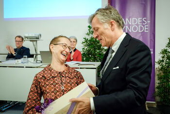 Eine weiblich zu lesende Person hält ein Paket in der Hand und schaut eine männlich zu lesende Person an. Beide lächeln.