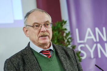 Eine männlich lesbare Person mit kurzen grauen Haaren und Brille am Rednerpult.