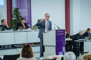 Eine männlich zu lesende Person steht an einem Redepult und spricht zu Menschen, die in Reihen an Tischen sitzen.