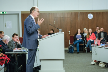Eine männlich zu lesende Person an einem Redepult vor sitzenden Menschen.