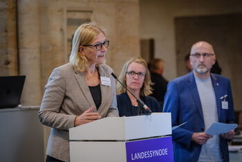 Eine Person steht an einem Rednerpult, in Hintergrund zwei weitere Personen