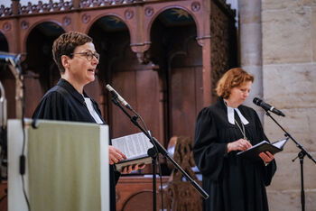 Zwei weiblich gelesene Personen mit Talar sprechen jeweils in ein Mikrofon. 