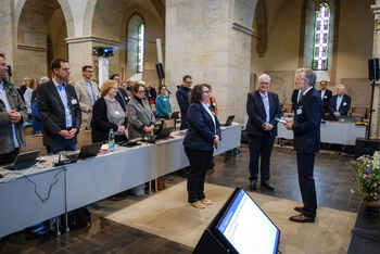 Eine Sitzung in einer Kirche, vorne stehen eine als Frau lesbare Person und zwei als Männer lesbare Personen.