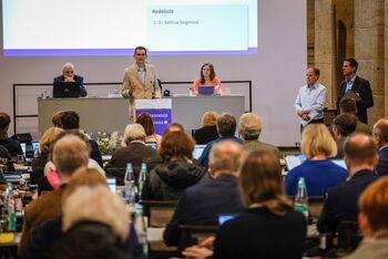 Plenumsteilnehmende sitzend im Vordergrund. Drei Personen im Fokus hinter einem Redepult.
