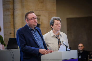 Eine weiblich und männlich lesbare Person stehen an einem Podium. Die männlich lesbare Person spricht.