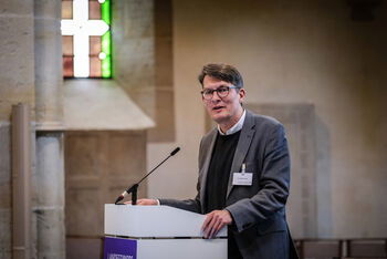 Eine männlich lesbare Person steht an einem Podium und spricht.