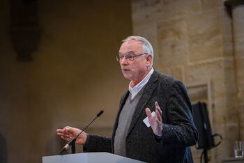 Eine männlich lesbare Person steht an einem Podium und spricht.