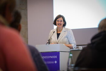 Eine als Frau lesbare Person steht an einem Redepult und spricht in ein Mikrofon.