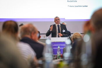 Eine männlich lesbare Person steht an einem Podium und spricht.