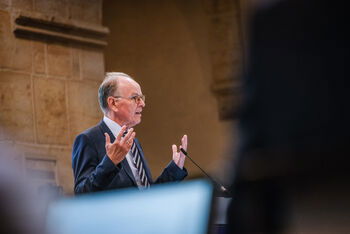 Eine männlich gelesene Person steht am Rednerpult und hebt beim Sprechen beide Hände. 