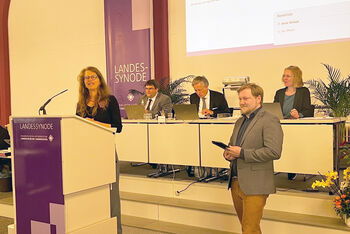 Eine weiblich zu lesende Person steht am Redepult und eine männlich zu lesende Person steht daneben.