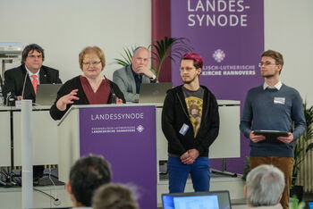 Eine Gruppe aus als Männer und Frauen lesbaren Personen an einem Redepult