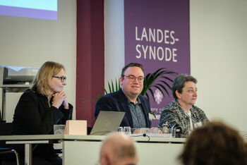 Eine männlich und zwei weiblich zu lesende Personen sitzen an einem Pult.