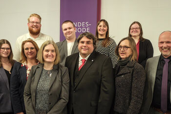 Sechs weiblich und vier männlich zu lesende Personen stehen als Gruppe zusammen und schauen in die Kamera.