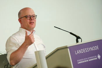 Eine männlich gelesene Person am Rednerpult im weißen Hemd und mit Brille. 