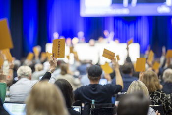 Menschen in einem blau beleuchteten Saal heben gelbe Karten zur Abstimmung.