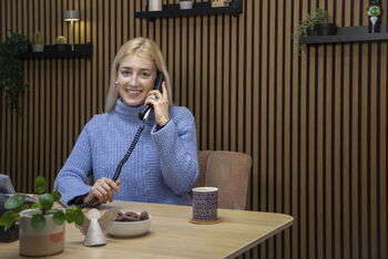 Eine als Frau lesbare Person sitzt an einem Tisch und telefoniert.