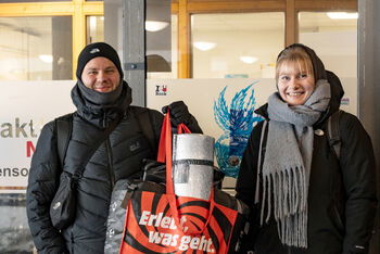 Eine als Mann und eine als Frau lesbare Person in Winterkleidung mit einem Beutel mit Schlafsack und Isomatte.
