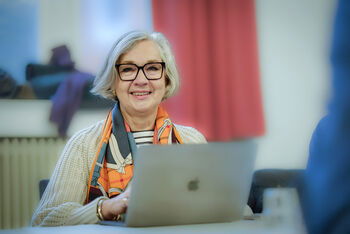 Eine weiblich gelesene Person mit mittellangen, grauen Haaren und buntem Halstuch sitzt an einem Laptop.