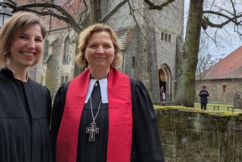 Zwei Frauen im Talar stehen vor einer Kirchenmauer.