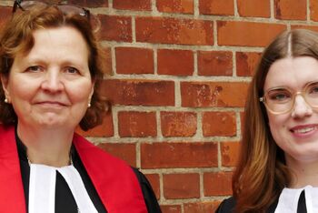 Zwei Frauen stehen im Talar vor einer Mauer.