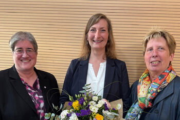 Drei als Frauen lesbare Personen, die mittlere hält einen Blumenstrauß.