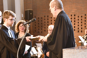 Mehrere Personen im Altarraum einer Kirche. Zwei männlich gelesene Personen in Talaren stehen sich gegenüber und überreichen eine Bibel.