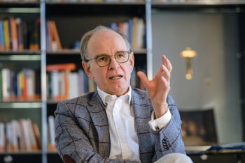 Eine als Mann lesbare Person mit Brille, weißem Hemd und Sakko sitzt vor einem Bücherregal, spricht und gestikuliert