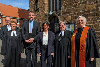 Eine Gruppe aus als Frauen und Männer lesbaren Personen, teils im Talar, vor einem Backsteingebäude.