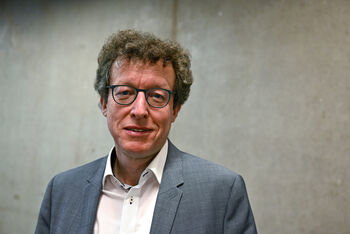 Eine als Mann lesbare Person mit Brille und Locken, steht im weißen Hemd und Sakko vor einer Betonwand.
