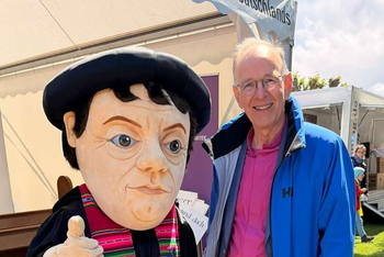 Eine männlich gelesene Person in blauer Jacke steht vor einem Zelt im Freien neben einer Figur, die Martin Luther ähnlich sieht. 