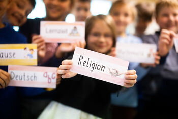 Kinder halten Schilder mit Schulfächern in die Kamera. Im Mittelpunkt steht Religion.