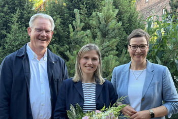 Zwei als Frauen lesbare Personen und eine als Mann lesbare Person posieren vor Gebüschen. Die Person in der Mitte hält einen Blumenstrauß in der Hand.