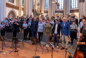 Mehrere als Frauen und Männer lesbare Personen stehen vor einem Orchester in einer Kirche.