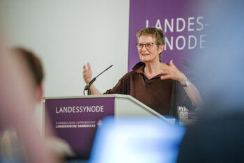 Eine als Frau lesbare Person steht an einem Redepuit und spricht gestenreich zu Publikum.