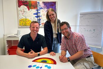 Drei lächelnde Personen sitzen an einem Tisch. Auf diesem Tisch liegt ein stilisierter Regenbogen. Im Hintergrund ist ein buntes Bild und eine Flipchart