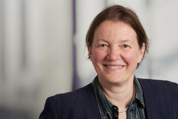 Portraitfoto einer als Frau lesbaren Person mit braunen zurückgebundenen Haaren.