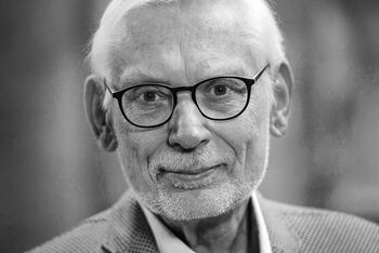 Eine männlich gelesene Person mit kurzen weißen Haaren, dunkler Brillenfassung und grauem Vollbart. Das Bild ist in schwarzweiß.