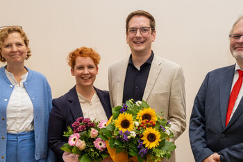 Vier Menschen, zwei als Männer, zwei als Frauen lesbar, stehen für ein Gruppenfoto. Die mittleren beiden halten Blumensträuße.