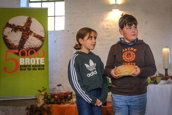 Eine als Mädchen und eine als Junge lesbare Person stehen mit einem Brot in einer Kirche neben einem Rollup mit der Aufschrift 5000 Brote.