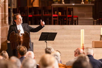 Eine als Mann lesbare Person im Anzug spricht vor Publikum in einer Kirche