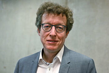 Eine als Mann lesbare Person mit Brille und Locken vor einer grauen Wand