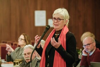 Eine als Frau lesbare Person mit rotem Schal spricht in ein Mikrofon.