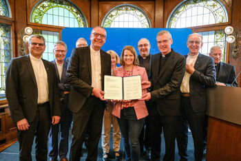 Ein Gruppe von weiblich und männlich zu lesenden Personen steht in einem Halbkreis und in der Mitte halten drei Personen ein Schriftstück in die Höhe.