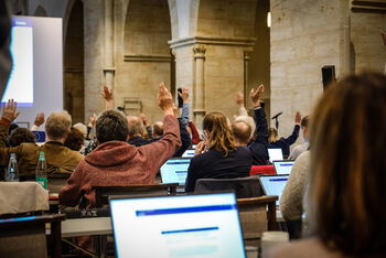Menschen sitzen in Reihen an Tischen in einer Kirche, manche haben Laptops vor sich. Einige heben die Hand zur Abstimmung.